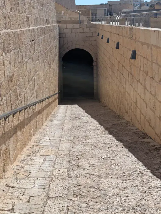 A sloping stone ramp leading up to a low vaulted tunnel entrance at Fort St Angelo, with the rampart wall rising on the left