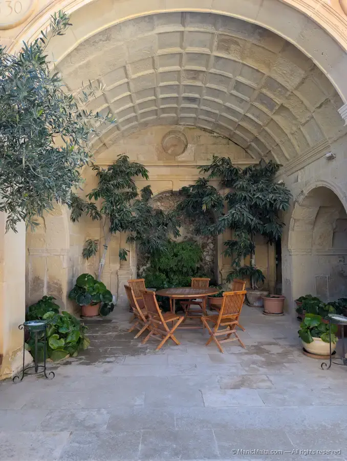 A small vaulted loggia at Fort St Angelo with olive trees in pots, a wooden table and chairs, set inside a stone alcove