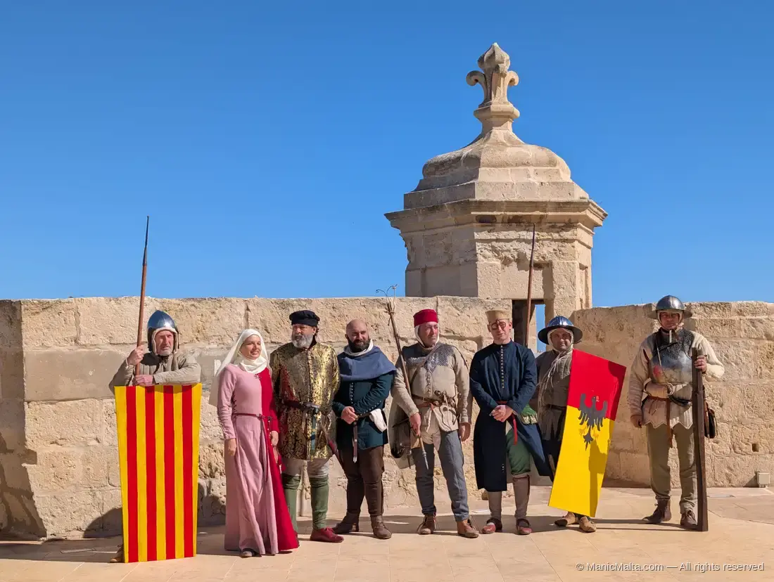 Group of seven costumed historical interpreters in Knights-era dress at Fort St Angelo, posed in front of a stone Vedette, with a pikeman, a noblewoman in a pink gown, knights, and a swordsman holding a striped heraldic shield