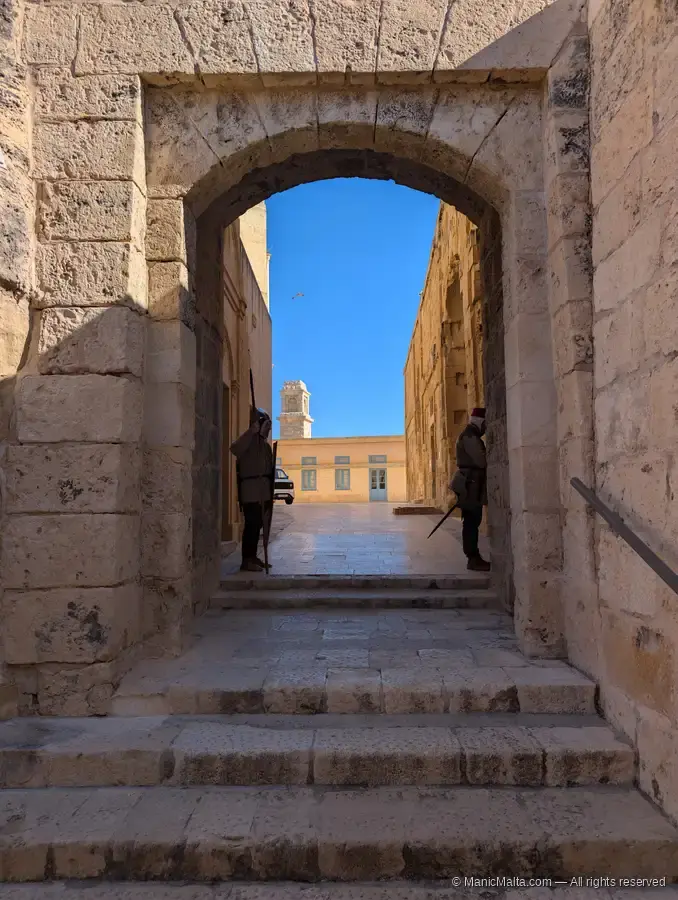 View through a high stone archway with steps inside Fort St Angelo, with two costumed soldier figures standing on either side