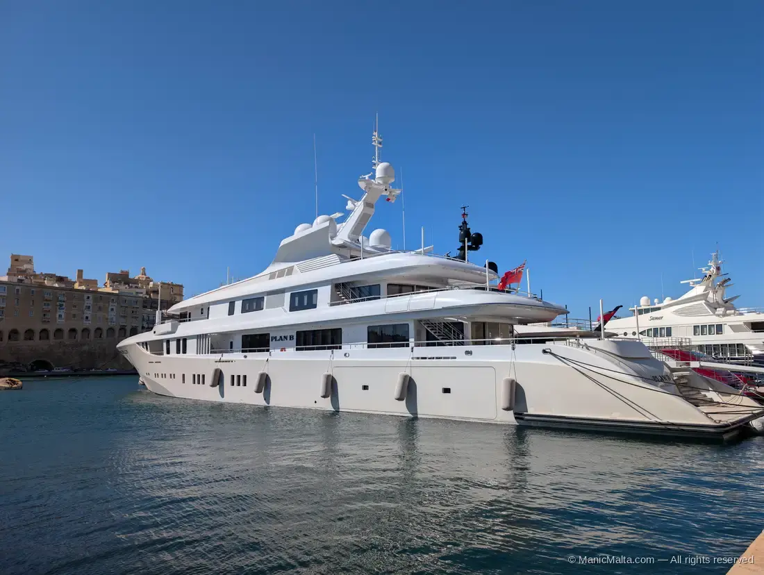 A large white superyacht moored at Vittoriosa marina directly beneath the stone walls of Fort St Angelo, with a smaller white yacht alongside