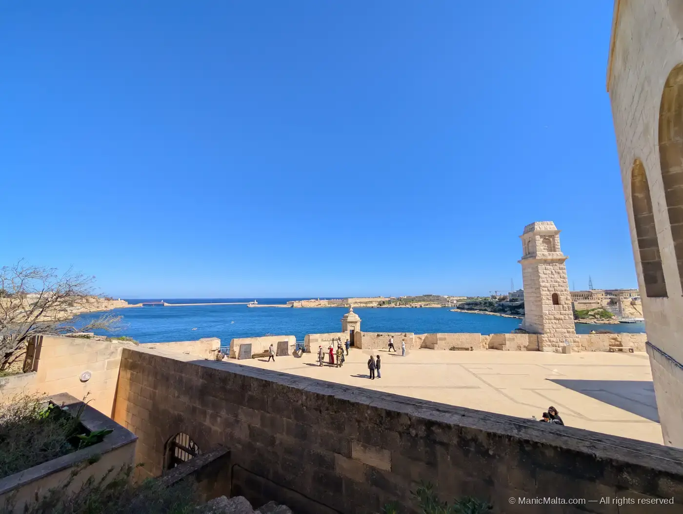Wide view from the upper terrace of Fort St Angelo across the Grand Harbour, with a small stone bell tower on the right, visitors walking on the plaza and the harbour mouth visible beyond