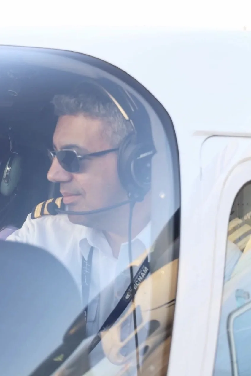 A Malta Wings pilot in uniform at the controls of the aircraft, seen through the cockpit window
