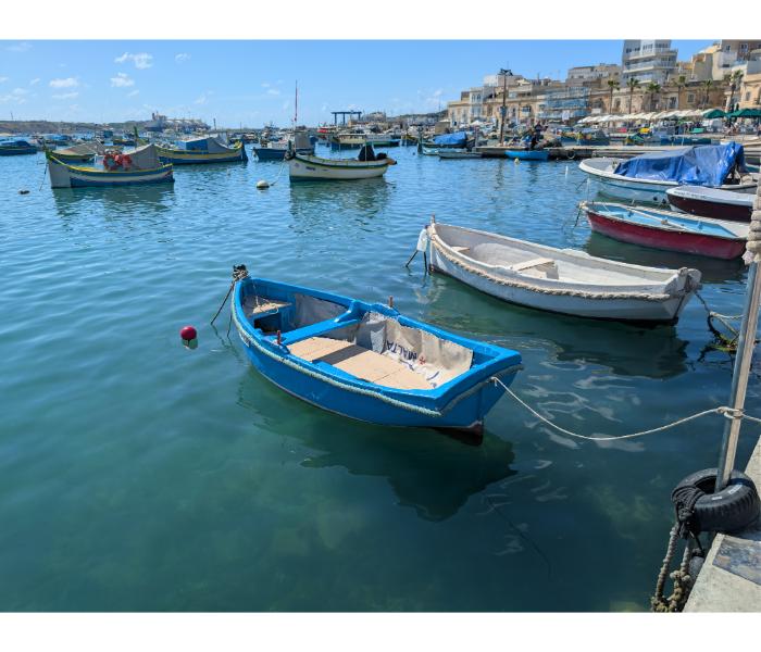 A small blue rowing boat in the foreground with the full sweep of Marsaxlokk harbour and its boats spread out behind