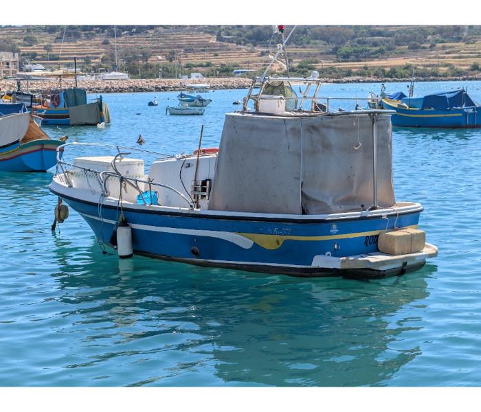 A blue and white motorboat moored at the Marsaxlokk quayside with a luzzu visible alongside and the village waterfront in the background