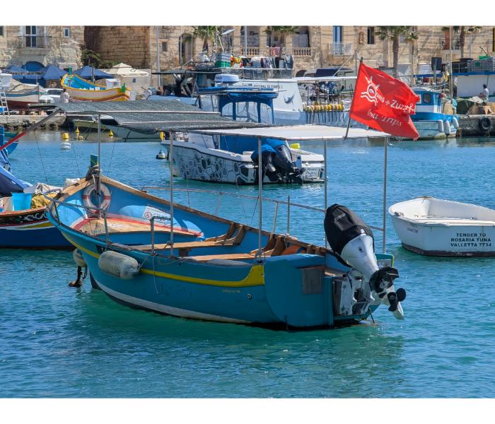Blue Captain Zuzu tour boat with a red banner flag and white sun canopy moored at the Marsaxlokk waterfront, with the village and a luzzu visible behind