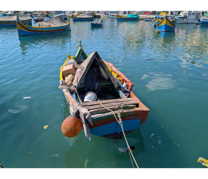 A small wooden rowing boat half-flooded and abandoned in Marsaxlokk harbour, photographed from above the quay with other boats moored in the background