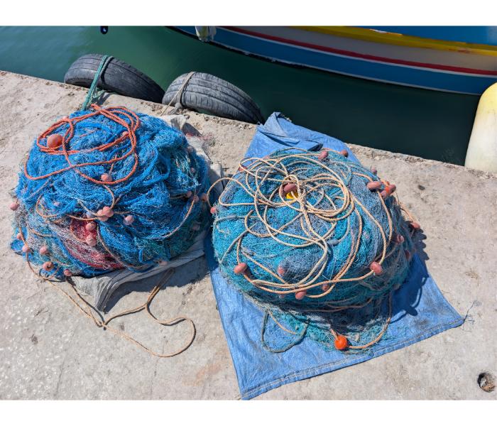 Two large piles of blue fishing nets with coiled rope and orange floats laid out on a tarp on the Marsaxlokk quay, with a luzzu hull visible behind