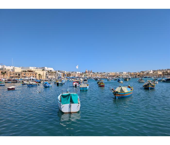 Wide panorama of Marsaxlokk harbour with the parish church of Our Lady of Pompei visible across the waterfront and dozens of small boats moored across the bay