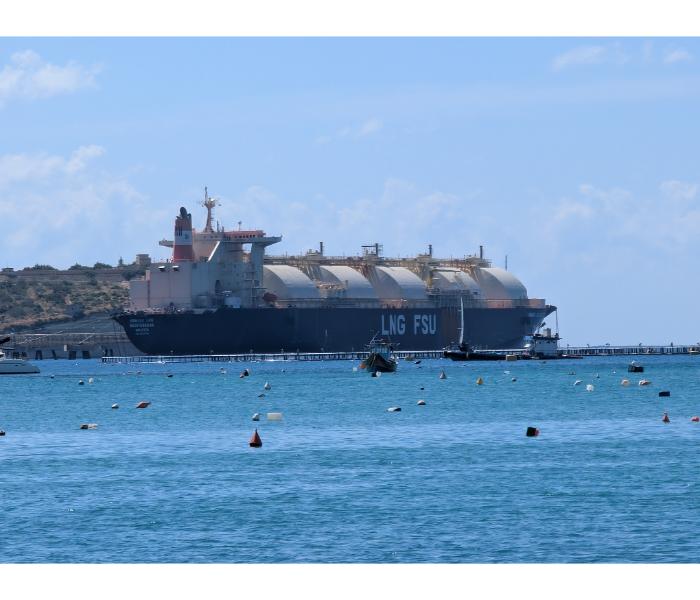 Side-on wide view of the LNG FSU floating storage unit moored in Marsaxlokk Bay, with its row of large spherical gas tanks visible along the deck and small mooring buoys in the foreground
