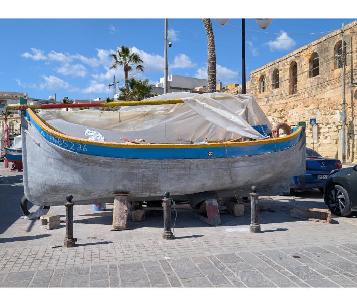 An unpainted Maltese fishing boat propped on stands ashore at Marsaxlokk with a canvas cover over the cockpit and a palm tree behind on the waterfront