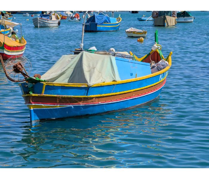 Maltese luzzu under a fitted canvas cover with bright yellow trim and traditional bow decoration, surrounded by other moored fishing boats in Marsaxlokk harbour