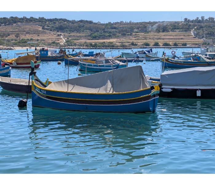 Yellow and blue luzzu numbered MFC 502 covered with a grey tarp, moored among other boats in Marsaxlokk harbour