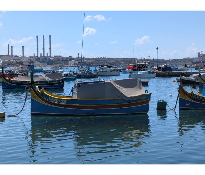 Two luzzu boats moored in Marsaxlokk harbour with the four red-and-white chimneys of Delimara power station rising directly behind them