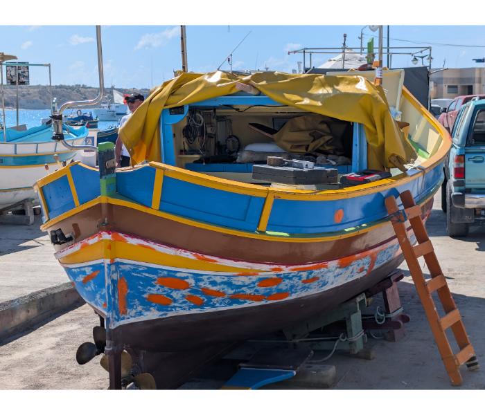 A luzzu mid-restoration ashore in Marsaxlokk with peeling paint on the hull, an orange ladder leaning against the side, and the owner working in the background