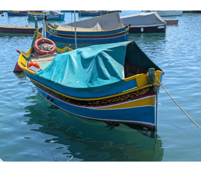 Maltese luzzu under a teal tarpaulin with bright yellow trim, floral painted decoration on the side, and the harbour quay behind