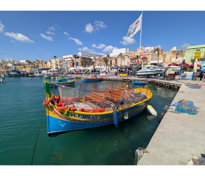 Luzzu VLT MFA 1297 in Marsaxlokk harbour with stacked wicker fish traps in the foreground and the Sunday market stalls being set up on the quay behind