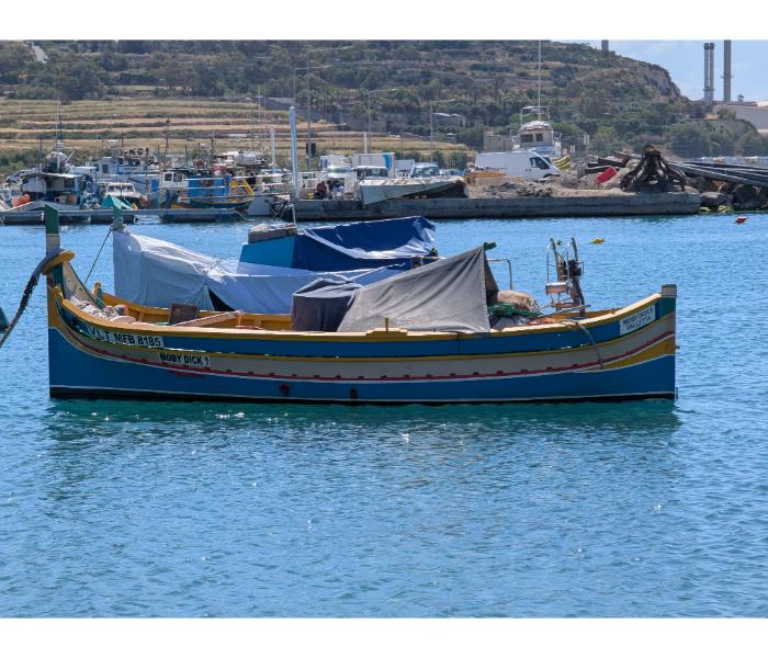 The luzzu Moby Dick 1 from Valletta with covered cockpit moored in Marsaxlokk with Delimara power station chimneys on the headland behind