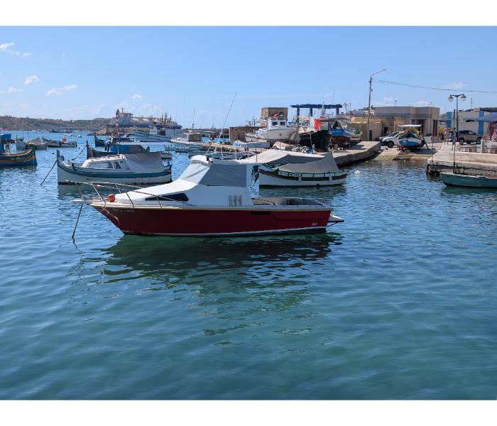 Red and white cabin cruiser at anchor in Marsaxlokk harbour with other fishing boats and the village waterfront visible behind