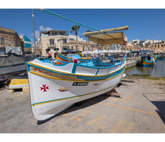 Restored white luzzu numbered S 24269 ashore in Marsaxlokk with the eight-pointed Maltese cross painted on the bow and the market crowd visible behind