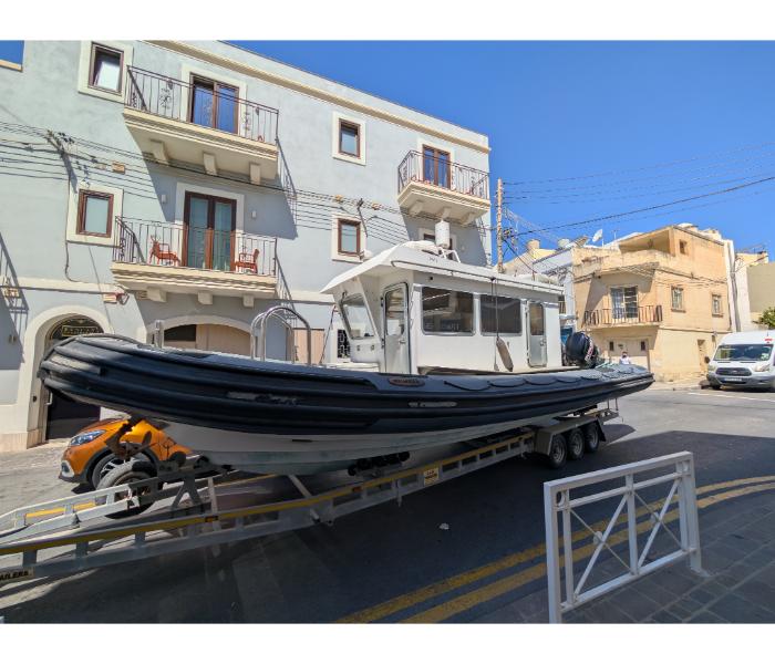 A large modern rigid inflatable boat on a trailer being towed through a narrow side street in Marsaxlokk village with traditional Maltese balconied houses on either side