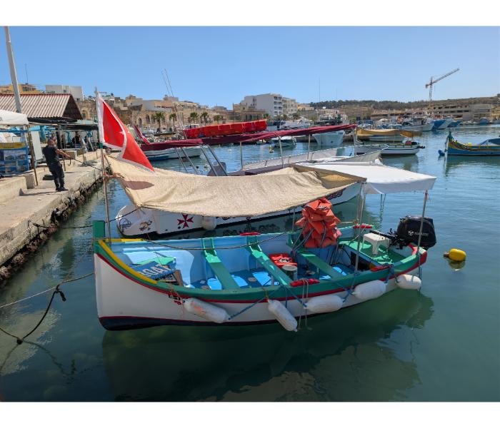 A traditional Maltese wooden rowing boat with a small Maltese cross flag, painted bench seats, and an orange figurehead, tied up at the Marsaxlokk waterfront