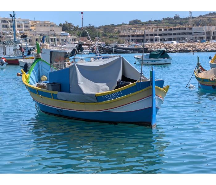 The luzzu San Pawl, painted blue and yellow with traditional Maltese trim, moored in Marsaxlokk harbour with a hillside village in the background