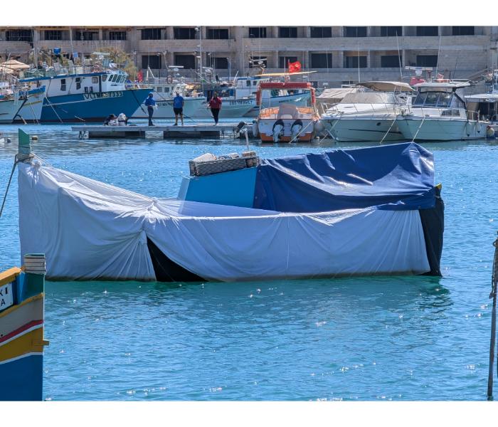 A boat almost entirely wrapped in a white tarpaulin floating in Marsaxlokk harbour, with the tarp tied down over what would be the cockpit