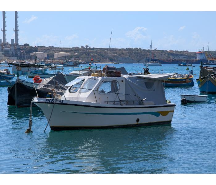 A small white cabin boat at the Marsaxlokk waterfront with the four red-and-white chimneys of Delimara power station rising directly behind on the headland