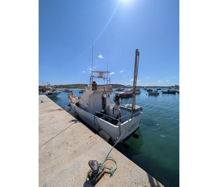 White motorboat moored at the Marsaxlokk stone quay on a still bright morning with the harbour and hillside visible across the bay