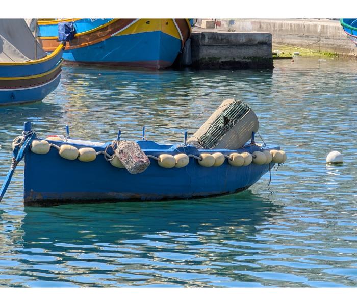 Blue Maltese workboat strung with a line of yellow round floats along the gunwale and a wicker fish trap on board, moored at the Marsaxlokk quay